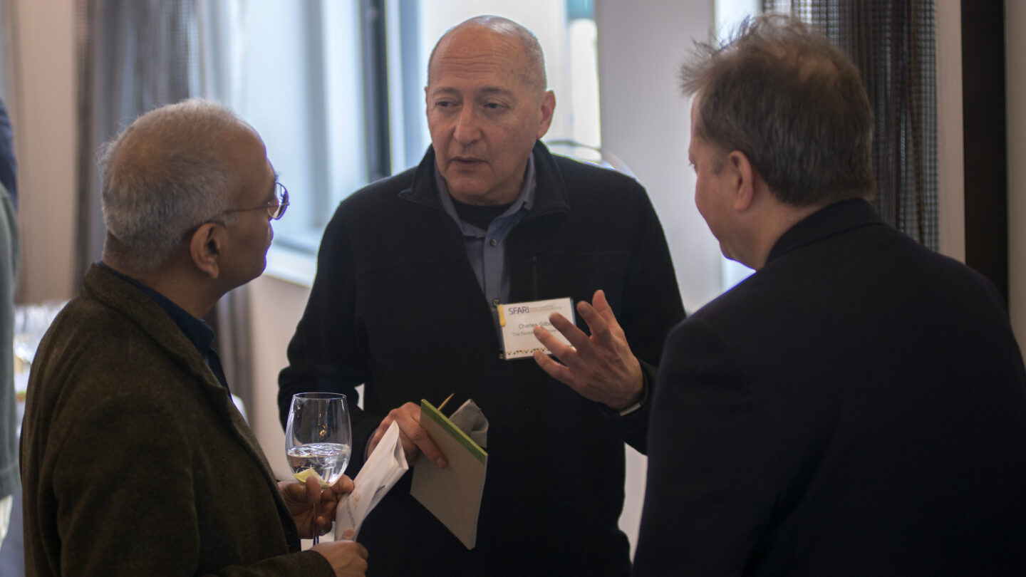 Three men stand indoors, engaged in conversation. One man holds a glass of water and paper, another gestures while speaking, and the third listens. They wear business casual attire and name tags.