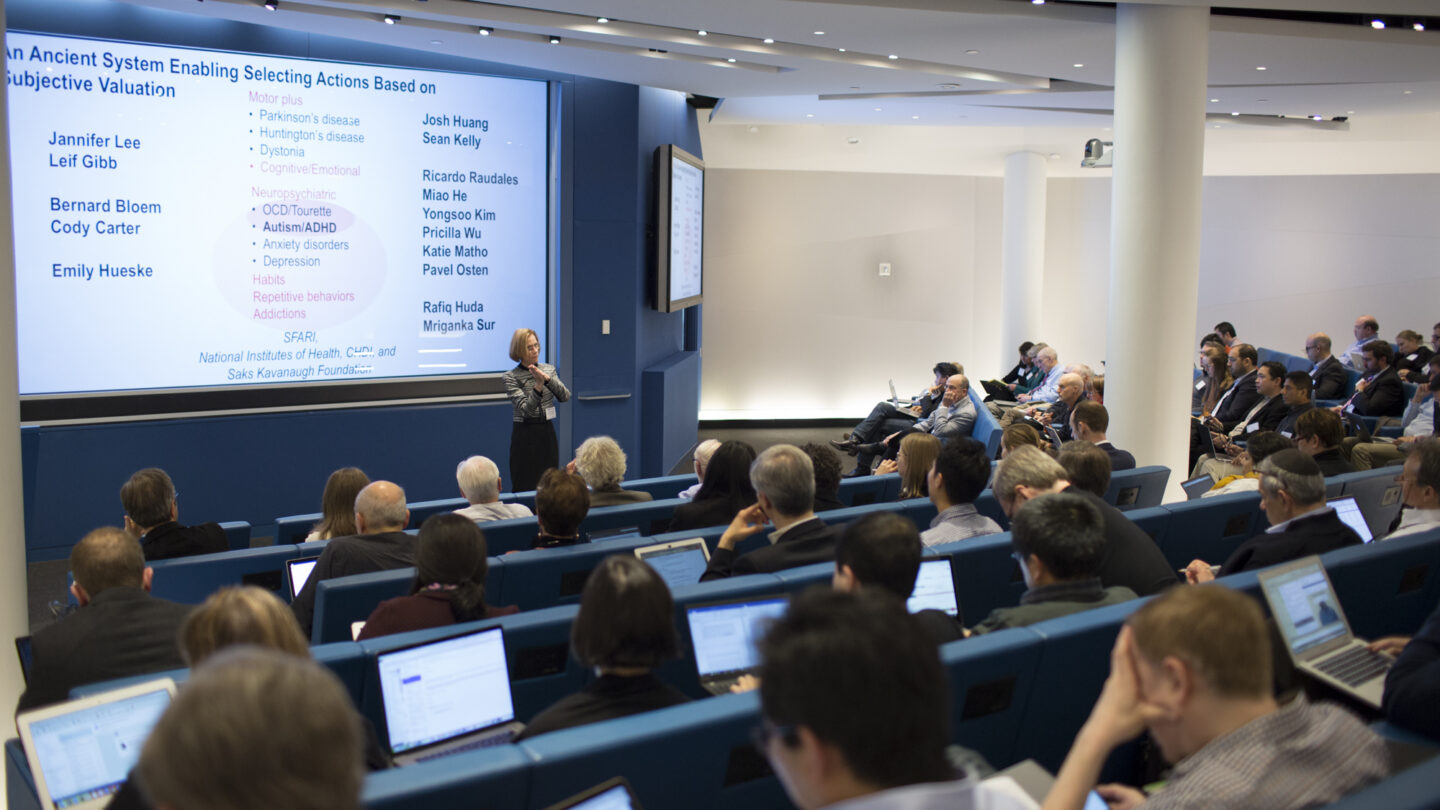A speaker presents slides in a modern lecture hall to an audience seated with laptops. Attendees listen, take notes, and the slide displays names and bullet points about an ancient system enabling actions.