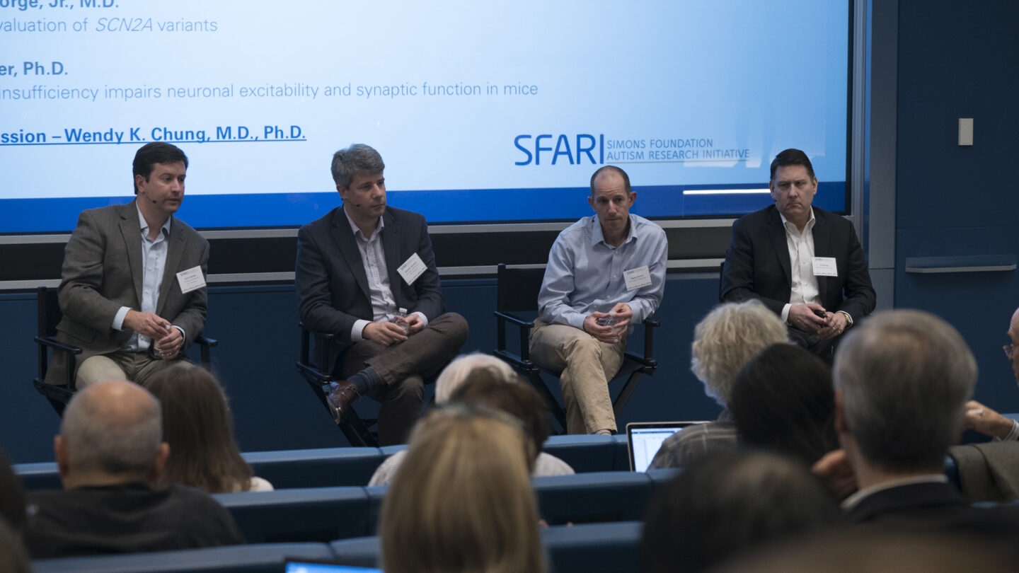 Four men in business attire sit on a panel in front of a blue screen, speaking to an audience in a lecture hall. The screen displays scientific text and the SFARI Simons Foundation Autism Research Initiative logo.