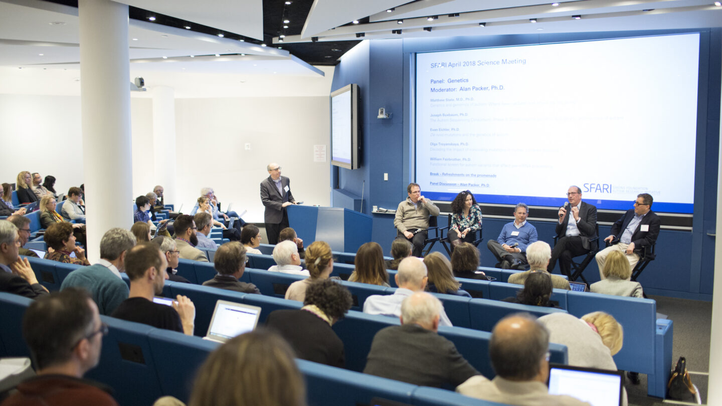 Audience members listen to a panel of five speakers on stage during a science meeting in a modern lecture hall with blue seating and large screen displays.