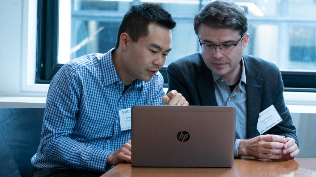 Two men sit at a table, closely looking at a laptop screen together. Both wear nametags and business attire, appearing engaged in discussion in a bright, modern office setting.