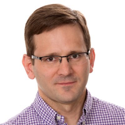 Man with short brown hair and glasses wearing a purple checkered shirt, looking at the camera with a neutral expression against a white background.
