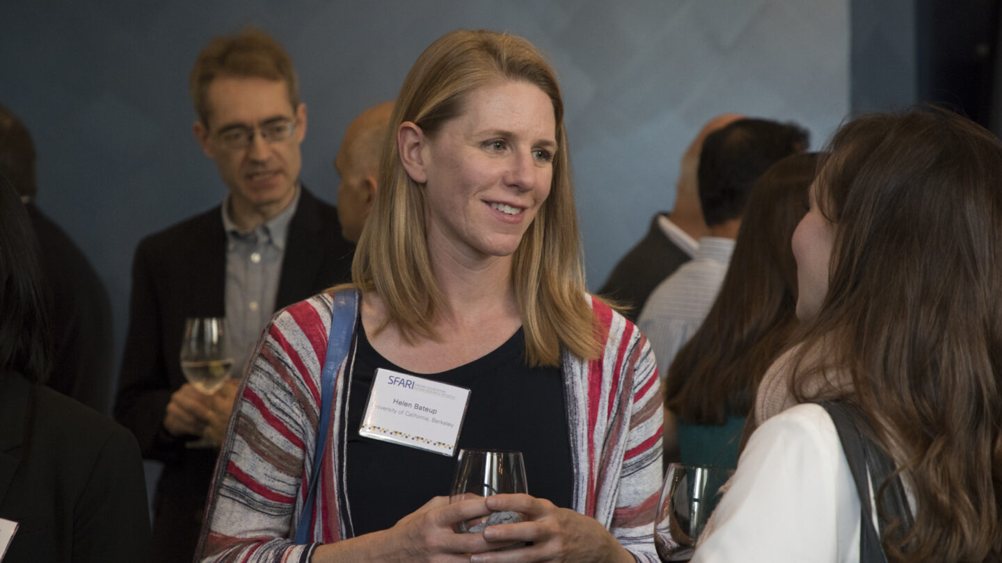 A woman with a name tag holding a wine glass talks to another woman at a networking event, with several people mingling in the background.