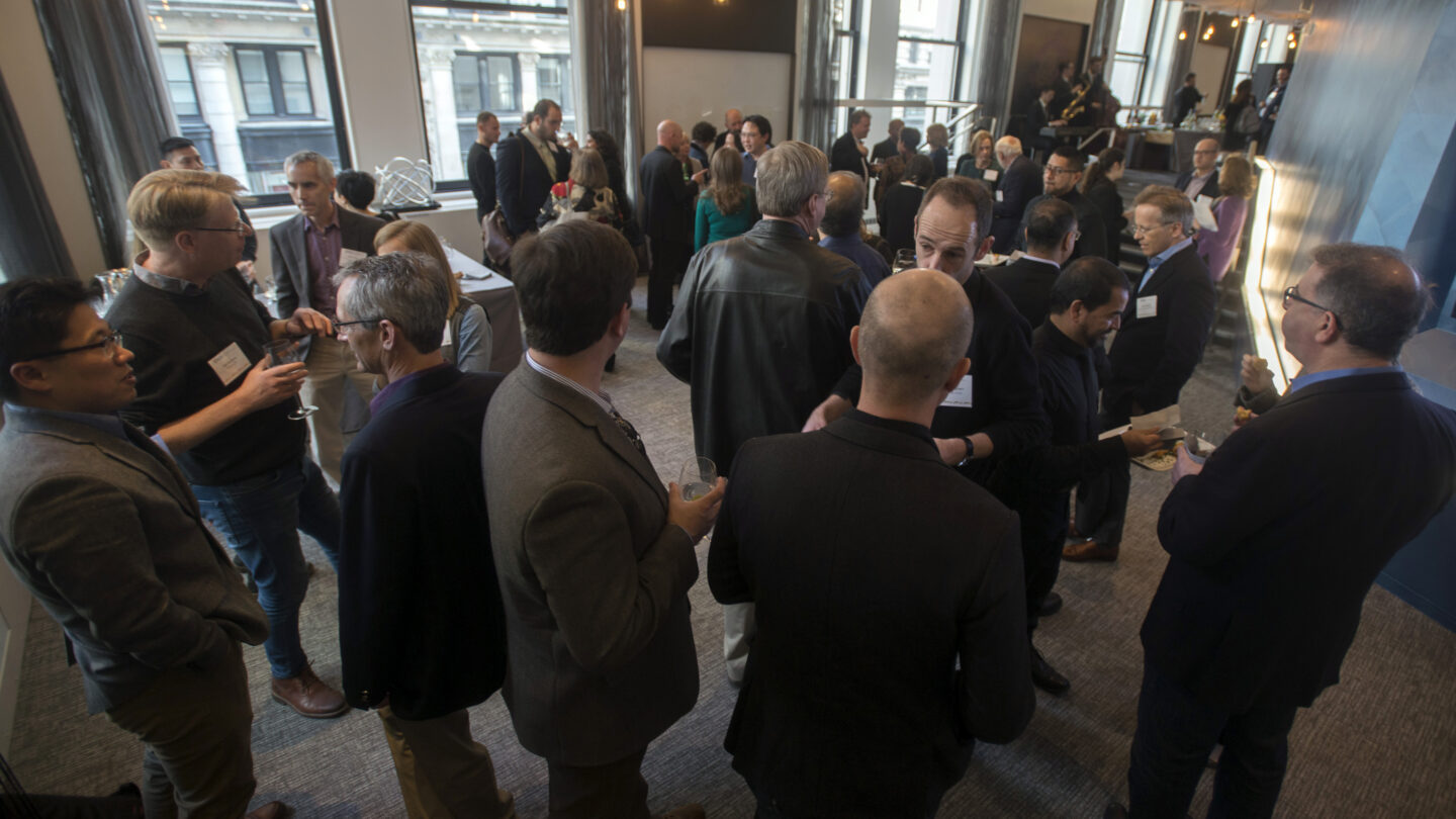 A group of people in business attire gather and mingle at a networking event in a modern, well-lit room with large windows and hanging lights.