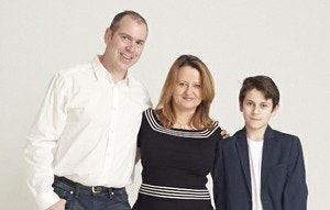 A man, woman, and boy stand together smiling. The man wears a white shirt and jeans, the woman wears a black dress, and the boy has on a dark blazer and white shirt, all against a plain light background.