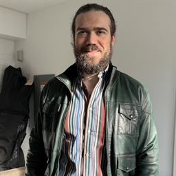 A man with a beard and long hair tied back is smiling. He is wearing a black leather jacket over a colorful striped shirt, standing indoors in natural light.