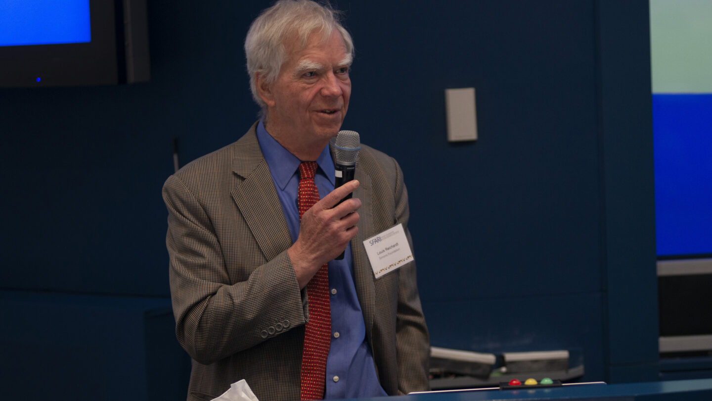 Older man with gray hair speaks into a microphone at a podium, wearing a suit and red tie, with a name tag visible. Blue background with screens and lights behind him.