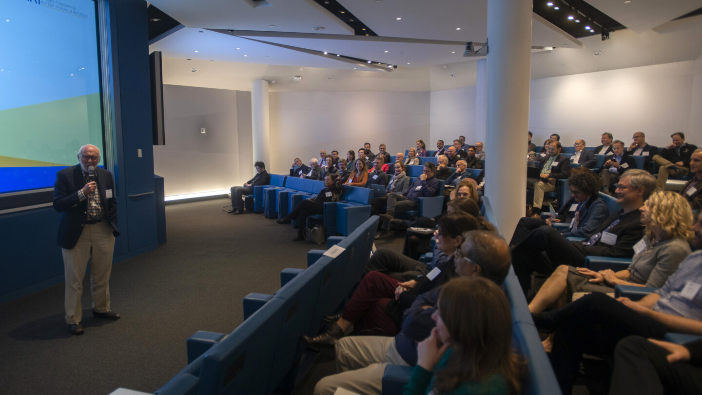 A man speaks at the front of a modern lecture hall filled with people seated in blue chairs, listening attentively. The large screen displays a colorful presentation.