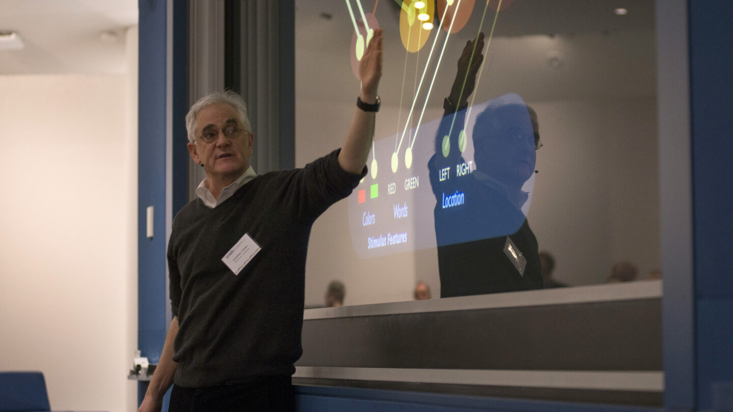 A man stands by a screen, pointing at a colorful flowchart with labeled circles and lines, while giving a presentation in a modern lecture room.