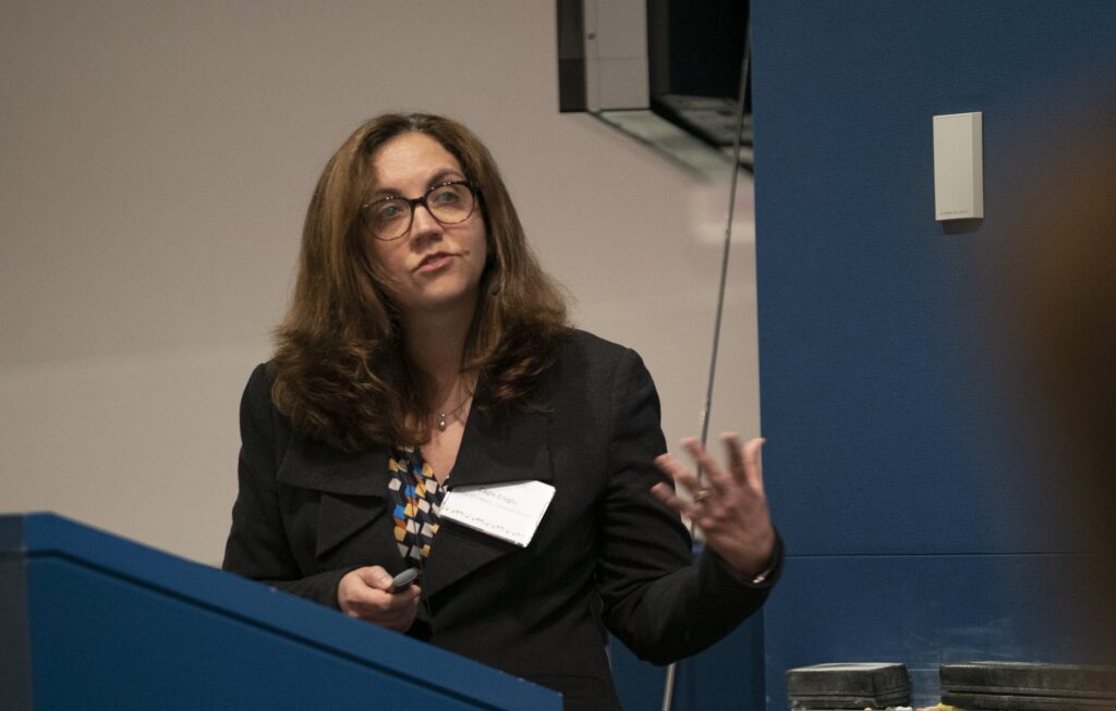 A woman with long brown hair and glasses speaks at a podium, gesturing with one hand. She is wearing a dark blazer and patterned blouse, and has a name tag on her chest. The background features a blue wall.