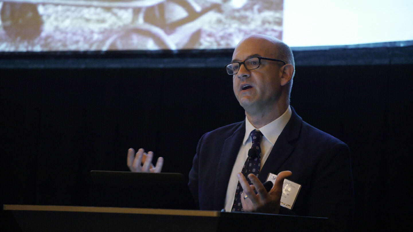 A man in a suit and glasses speaks at a podium, gesturing with both hands. A large screen with an image is visible behind him.