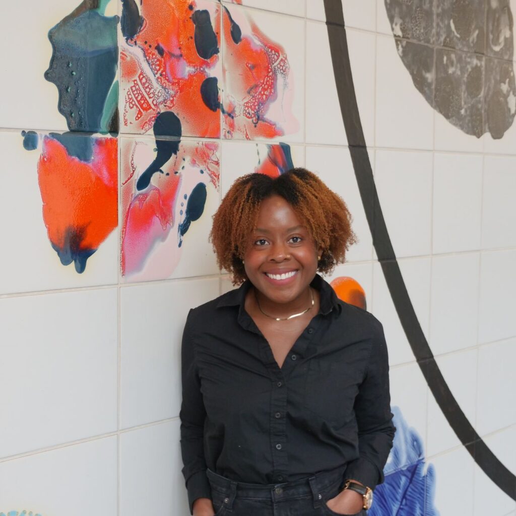 A person with short curly hair, wearing a black shirt and pants, stands smiling in front of a white tiled wall decorated with colorful abstract art.