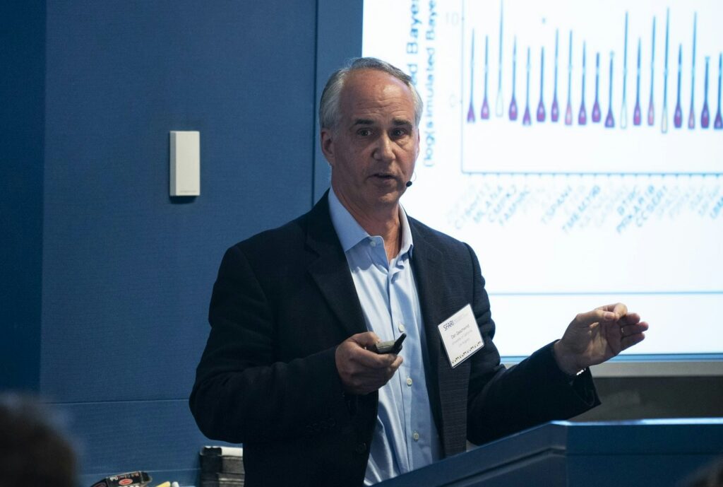 A man in a suit gives a presentation, speaking into a headset microphone, with a graph displayed on a screen behind him.