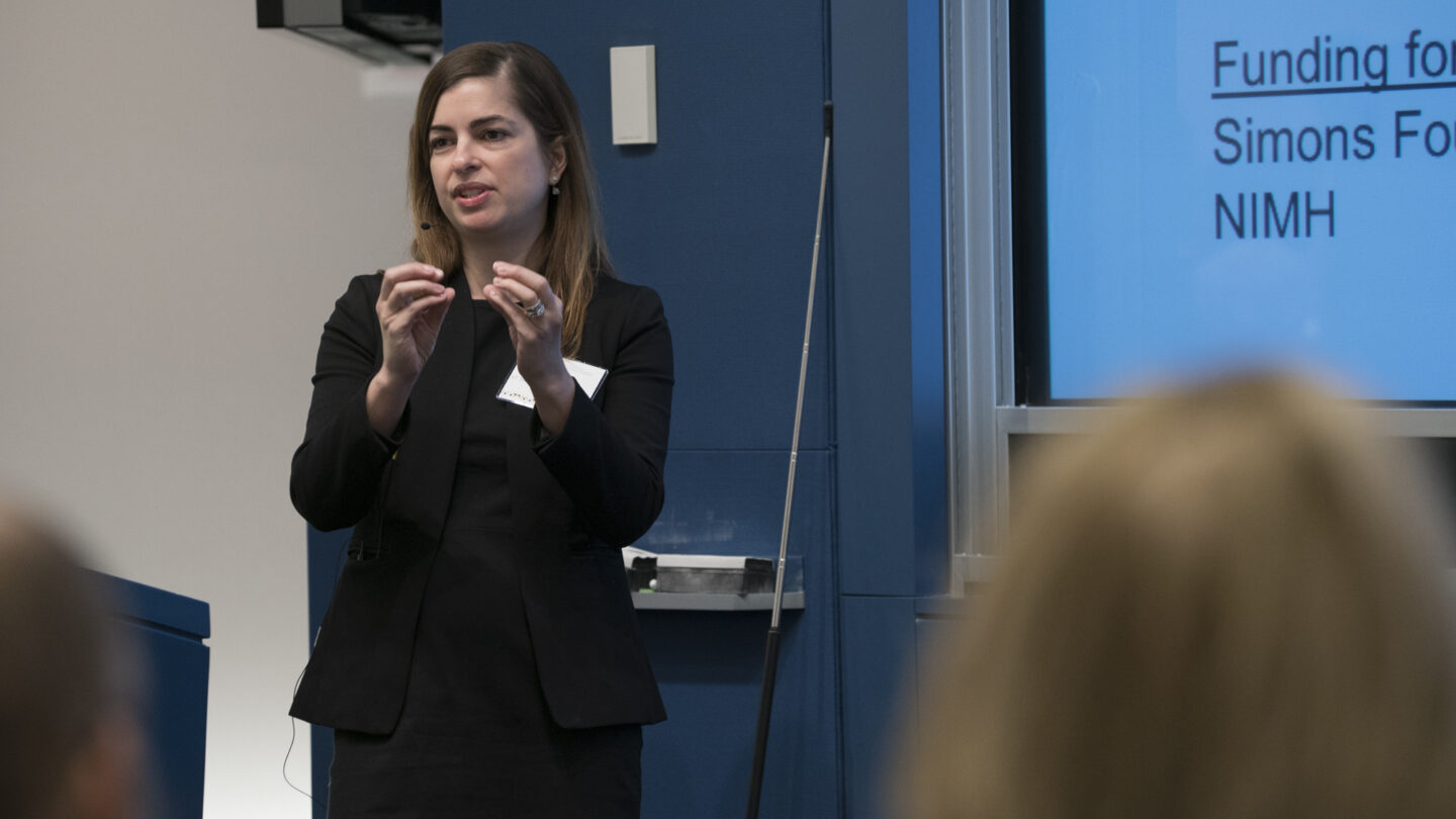 A woman in a black suit speaks and gestures in front of an audience, with a presentation slide about funding from the Simons Foundation and NIMH visible on the screen beside her.