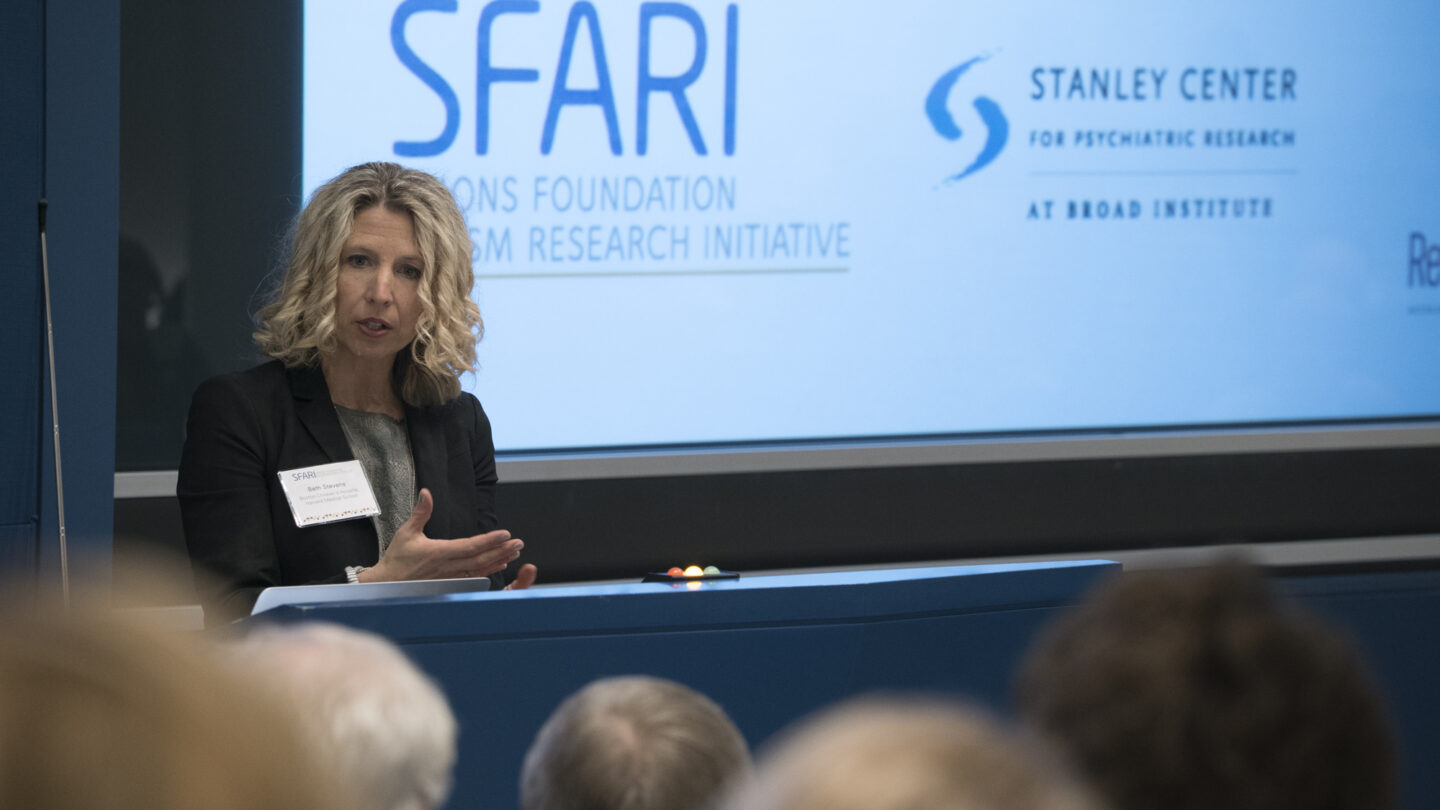 A woman speaks at a podium during a conference. Behind her, screens display the SFARI logo and the Stanley Center for Psychiatric Research logo. Audience members are visible in the foreground.