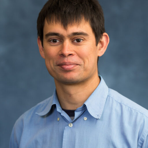 A man in a light blue button-up shirt stands in front of a mottled blue background, looking at the camera with a slight smile.