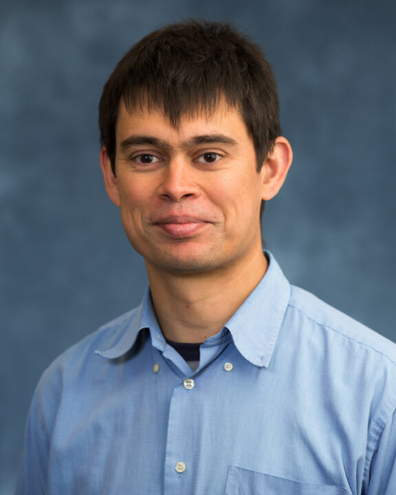 A man in a light blue button-up shirt stands in front of a mottled blue background, looking at the camera with a slight smile.