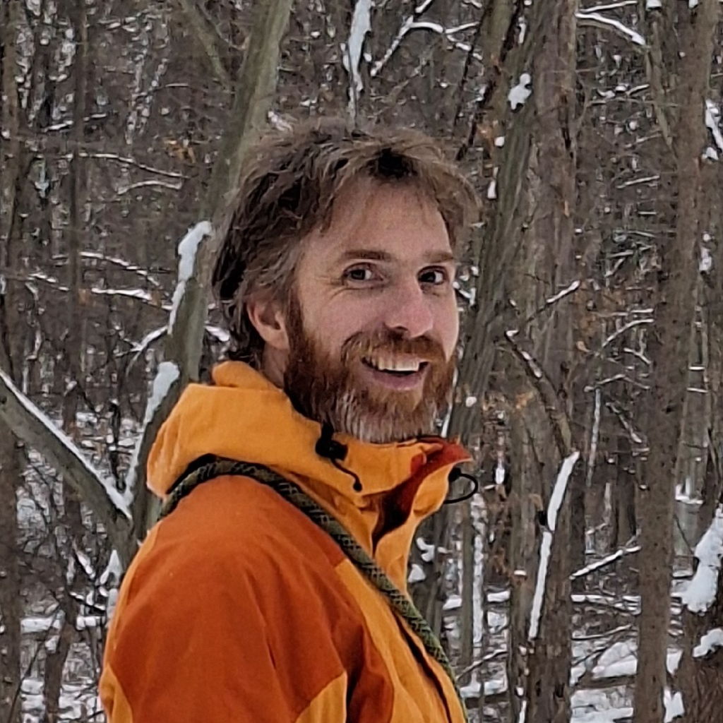 A man in an orange jacket stands in a snowy forest, smiling at the camera. The background is filled with leafless trees and snow.