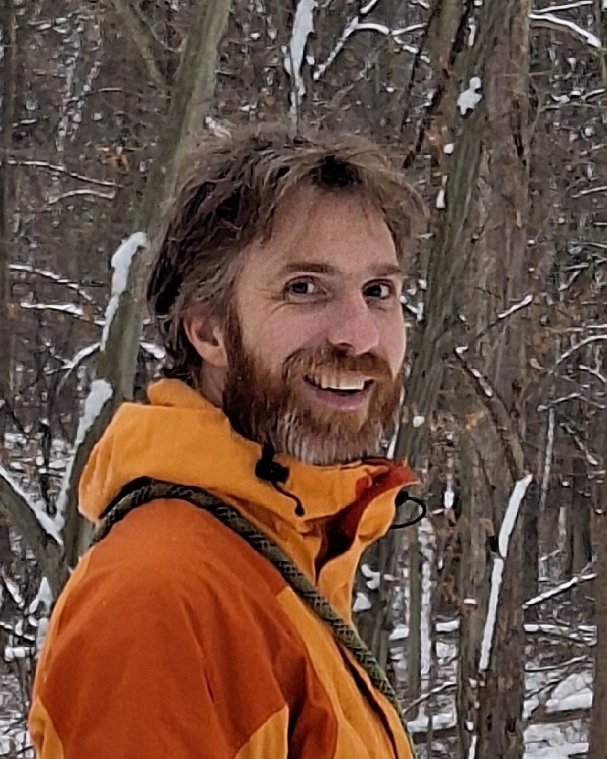 A man in an orange jacket stands in a snowy forest, smiling at the camera. The background is filled with leafless trees and snow.