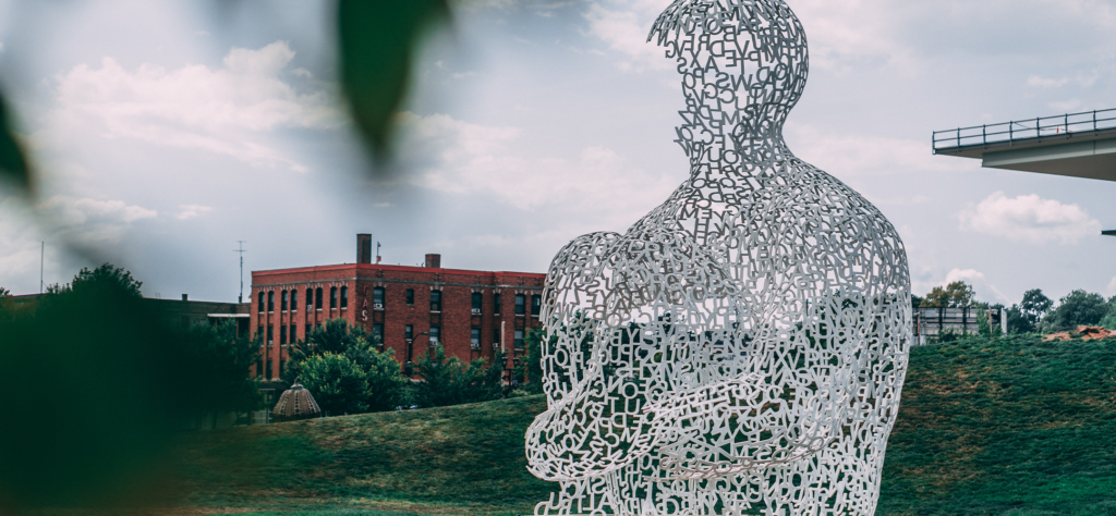 A large sculpture of a human figure made from white letters stands on grass, with a red brick building and trees in the background under a partly cloudy sky.