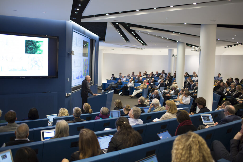 A speaker presents scientific data to a large, attentive audience in a modern lecture hall. Attendees sit in blue chairs, many using laptops, as graphs and images are displayed on big screens.