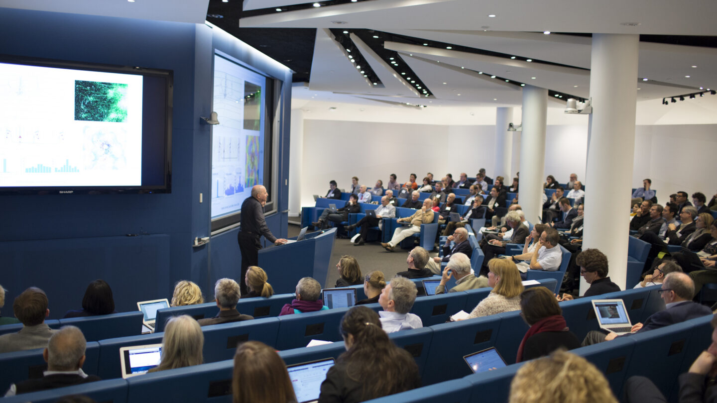 A speaker presents scientific data to a large, attentive audience in a modern lecture hall. Attendees sit in blue chairs, many using laptops, as graphs and images are displayed on big screens.