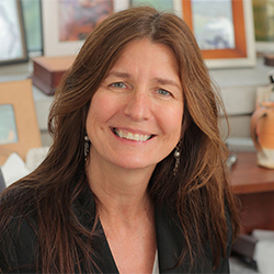 Smiling woman with long brown hair sits indoors, wearing a dark blazer. Behind her are framed pictures and a ceramic vase on a shelf, giving the setting a warm and personal feel.