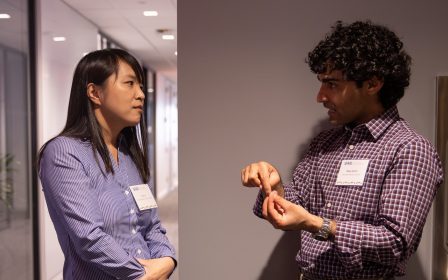 Two people stand in a hallway, facing each other and talking. Both wear name tags and business casual clothes. The man gestures with his hands while the woman listens with her arms folded.