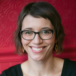 A woman with short brown hair and glasses smiles at the camera. She is wearing earrings and a black top, with a textured red wall in the background.