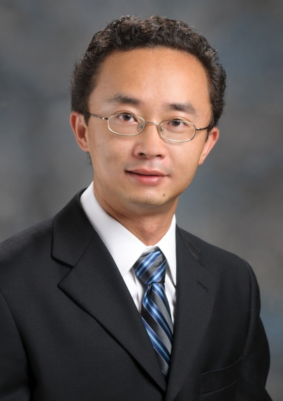 A man wearing glasses, a black suit, a white shirt, and a blue striped tie poses for a professional portrait against a gray background.