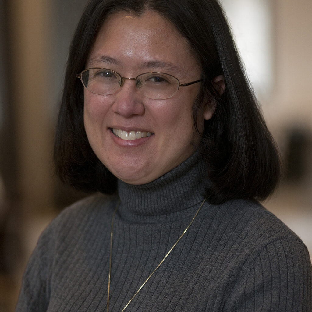 A woman with shoulder-length dark hair and glasses smiles while wearing a gray ribbed turtleneck sweater and a gold necklace. The background is softly blurred.