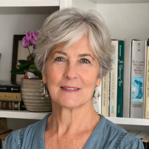 An older woman with short gray hair and earrings, wearing a blue top, stands in front of a bookshelf with books and a potted plant. She is smiling slightly at the camera.