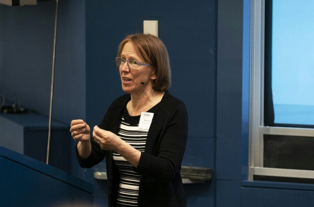 A woman wearing glasses and a black cardigan speaks at a podium in a blue room, gesturing with her hands. A name tag is visible on her chest, and she wears a headset microphone.