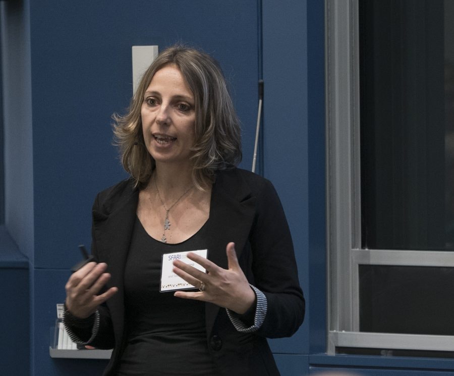 A woman with shoulder-length brown hair speaks while gesturing with her hands in a room with blue walls and a window. She wears a black blazer and a necklace, and has a name tag on her chest.