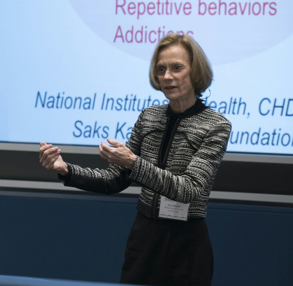 A woman in glasses and a striped jacket is speaking and gesturing in front of a presentation slide featuring text about health, behaviors, and foundations.