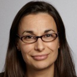 A woman with long brown hair and glasses smiles slightly, facing the camera against a plain light background.