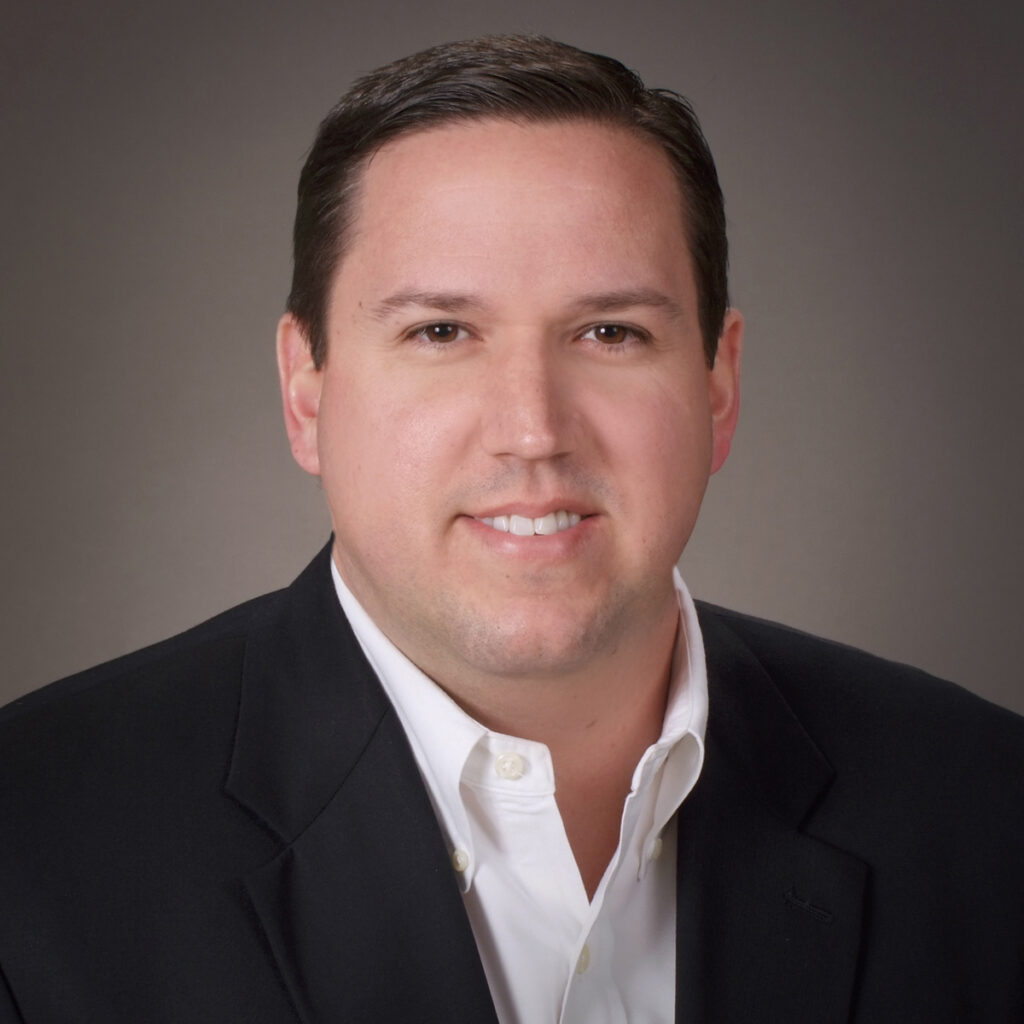 A man with short dark hair in a black blazer and white shirt smiles at the camera against a plain, gray background.