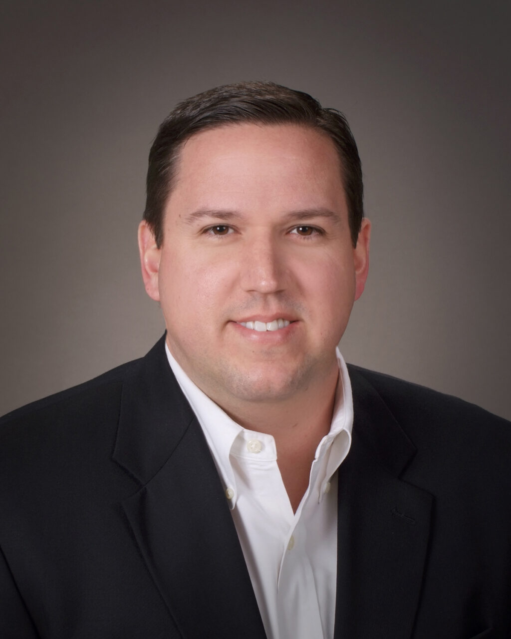 A man with short dark hair in a black blazer and white shirt smiles at the camera against a plain, gray background.