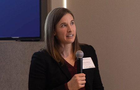 Woman with long brown hair holding a microphone, wearing a black blazer and name tag, speaking in front of a screen in a conference room.