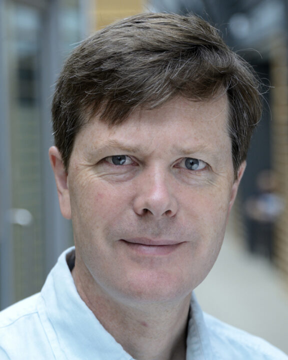 A man with short brown hair and blue eyes, wearing a light blue collared shirt, stands indoors with a neutral expression. The background is blurred with glass and wooden elements.