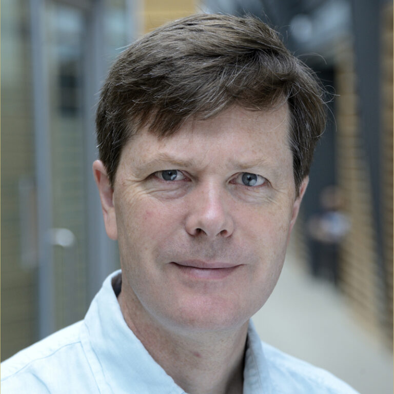 A man with short brown hair and blue eyes, wearing a light blue collared shirt, stands indoors with a neutral expression. The background is blurred with glass and wooden elements.