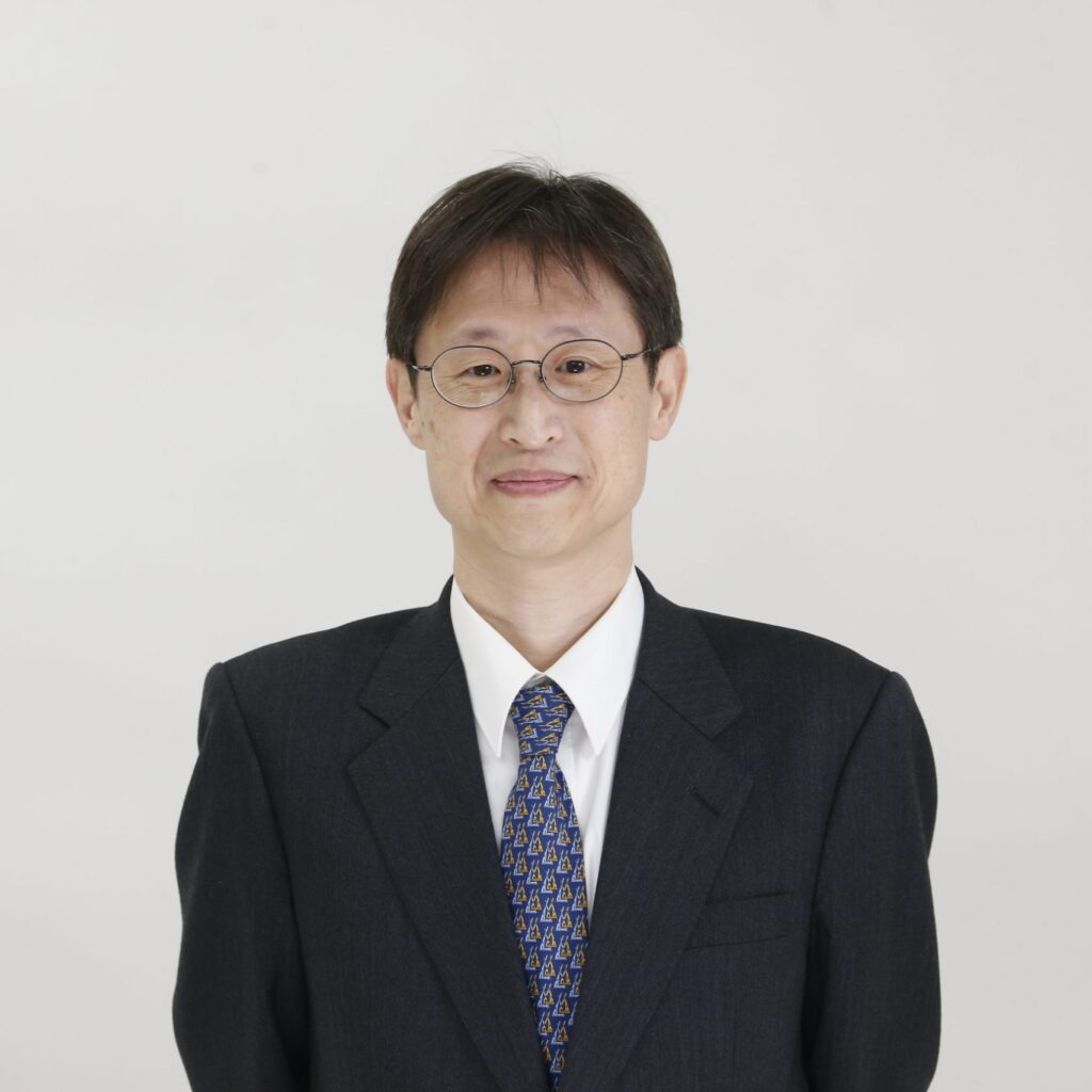 A man wearing glasses, a dark suit, and a patterned tie stands against a plain white background, smiling softly at the camera.