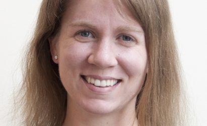 A woman with straight light brown hair, blue eyes, and fair skin smiles at the camera against a plain light background.