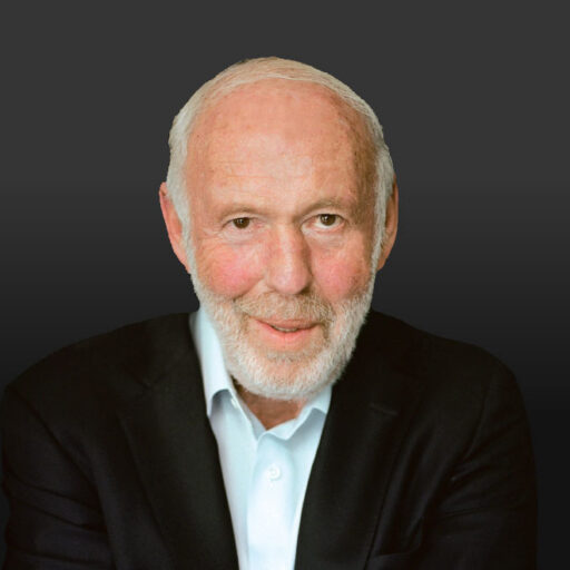 An older man with a white beard and short hair wears a dark suit jacket and light blue shirt, smiling slightly against a plain dark background.