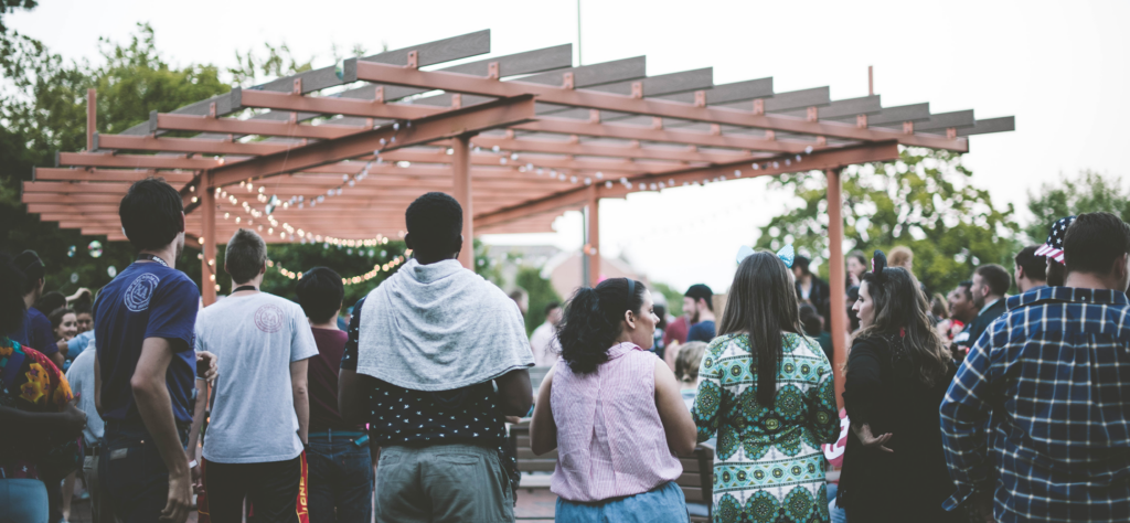 A group of people seen from behind stand together under a wooden pergola decorated with string lights.