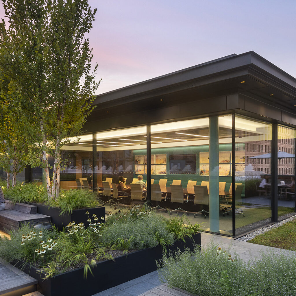 People sit at tables on a rooftop patio with trees and planters, next to a modern glass-walled meeting room. City buildings and a golden-domed structure are visible in the background at sunset.