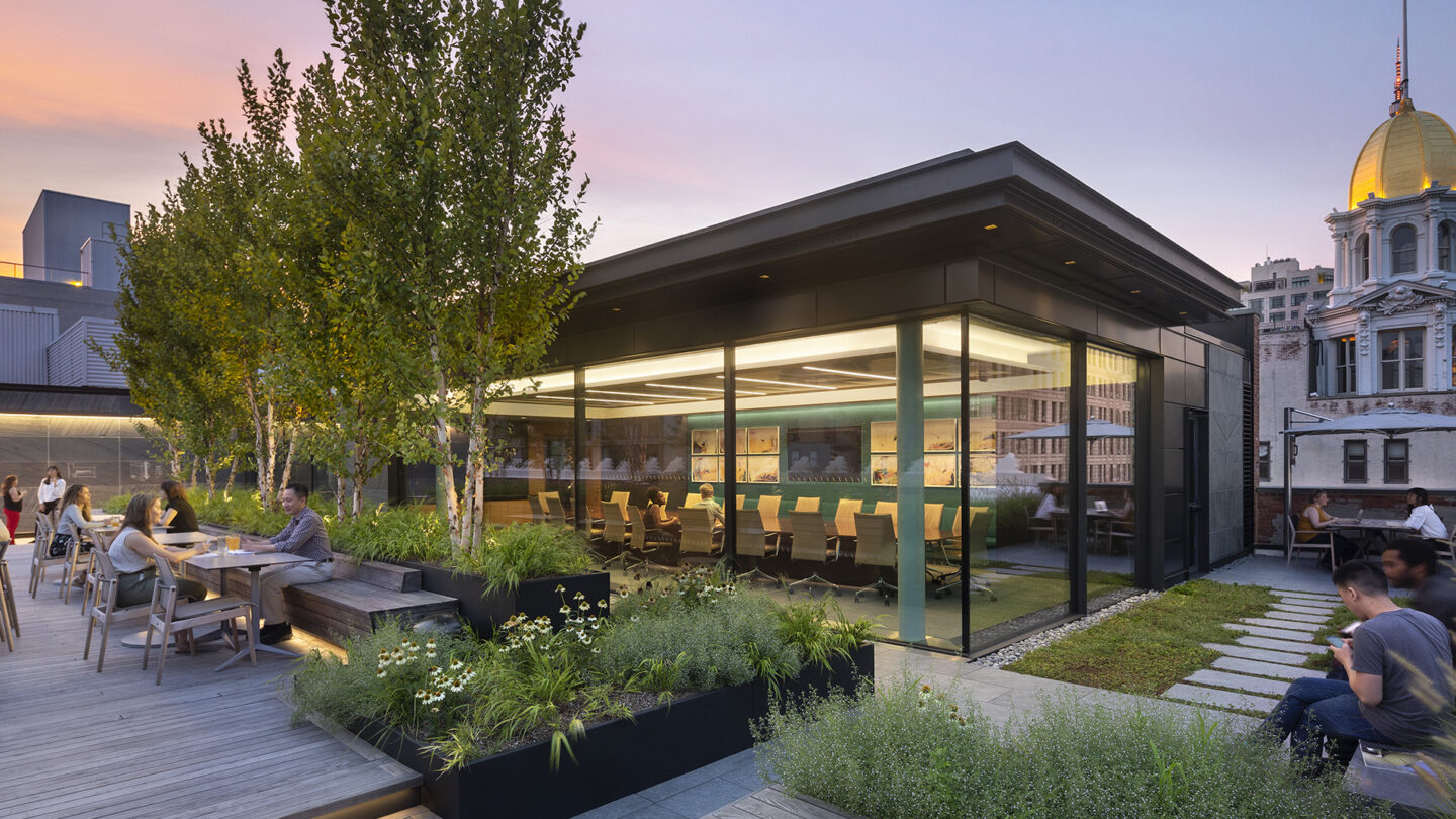 People sit at tables on a rooftop patio with trees and planters, next to a modern glass-walled meeting room. City buildings and a golden-domed structure are visible in the background at sunset.