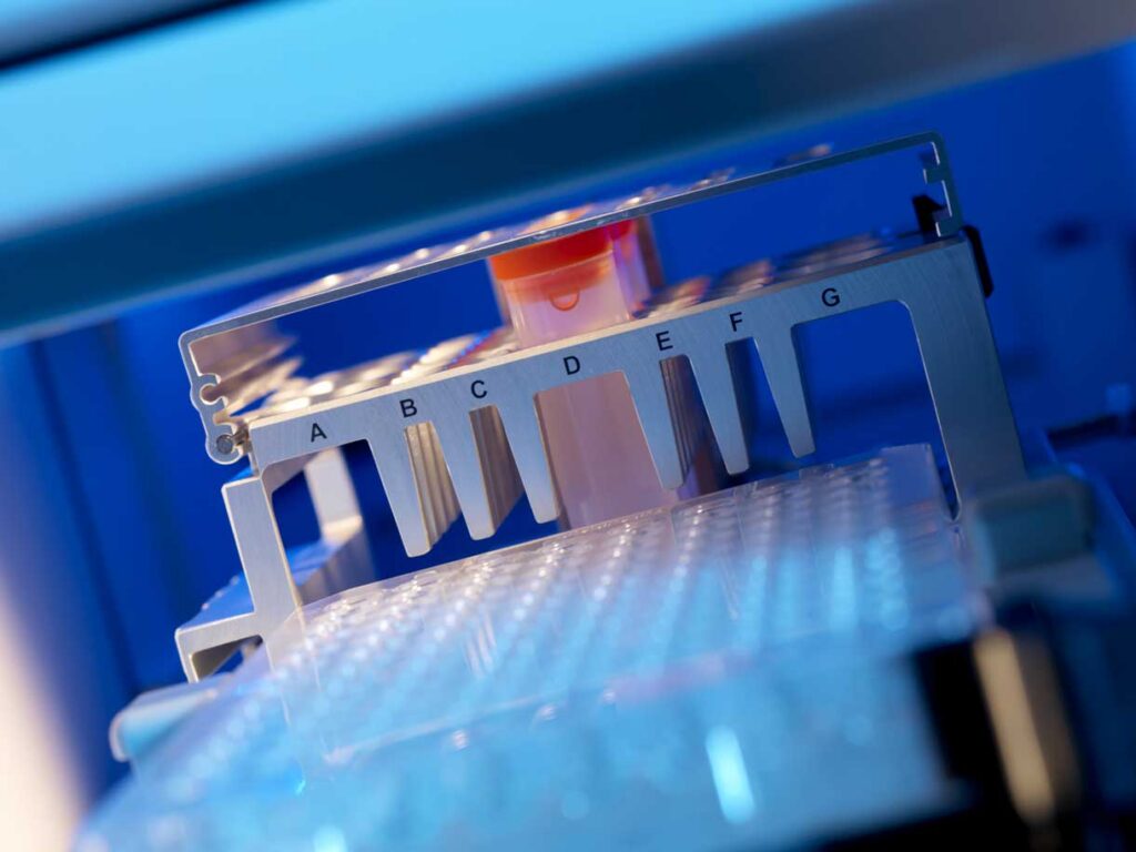 Close-up of a laboratory automated pipetting system with labeled slots A–G and a single test tube inserted, illuminated by blue light.