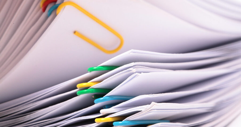 A close-up view of several stacks of papers clipped together with colorful paperclips in yellow, green, and blue. The image focuses on the edges and corners of the documents.