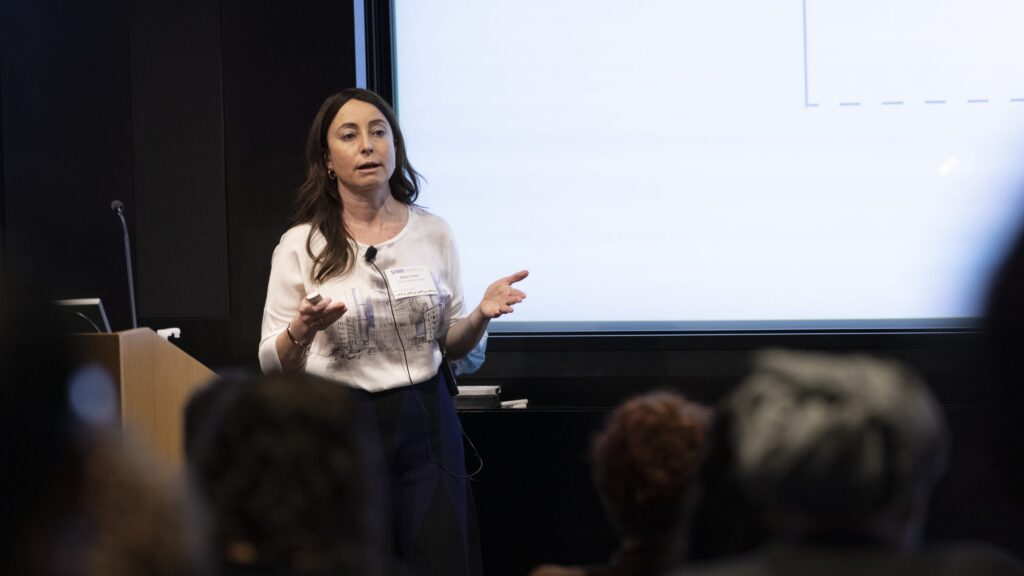A woman stands in front of a screen giving a presentation to an audience, gesturing with her hands.
